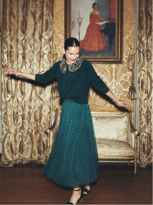 Woman modelling a dark green knit jumper with embellished collar and teal pleated midi skirt in an elegant vintage interior.