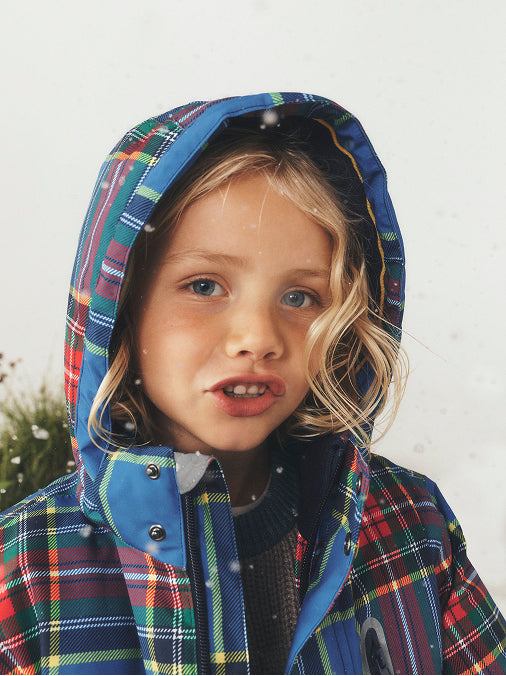 A young child wearing a hooded plaid jacket with blue trim, photographed outdoors against a light neutral background with snowflakes falling.