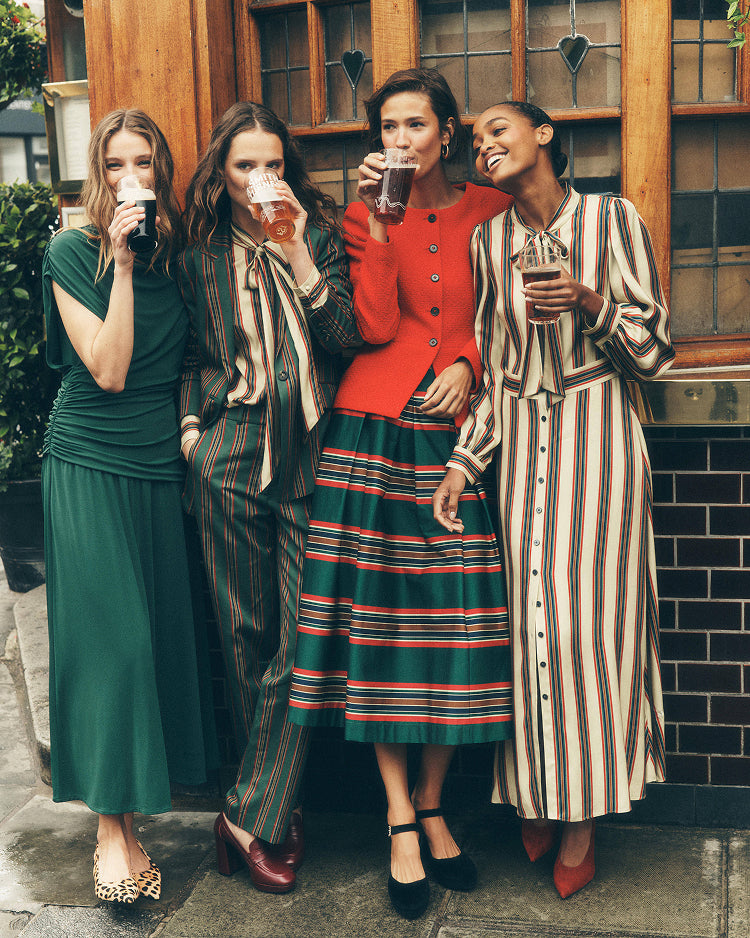 Four women standing together outdoors in front of a wooden building, each holding a drink. They are wearing stylish coordinated outfits, including green, striped beige-and-brown, and red-and-green striped garments.