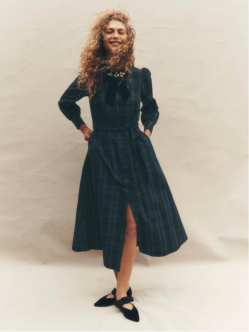 A woman modeling a dark teal plaid midi dress with long sleeves, a high-low skirt, and a front slit, paired with black flats, against a neutral beige backdrop.