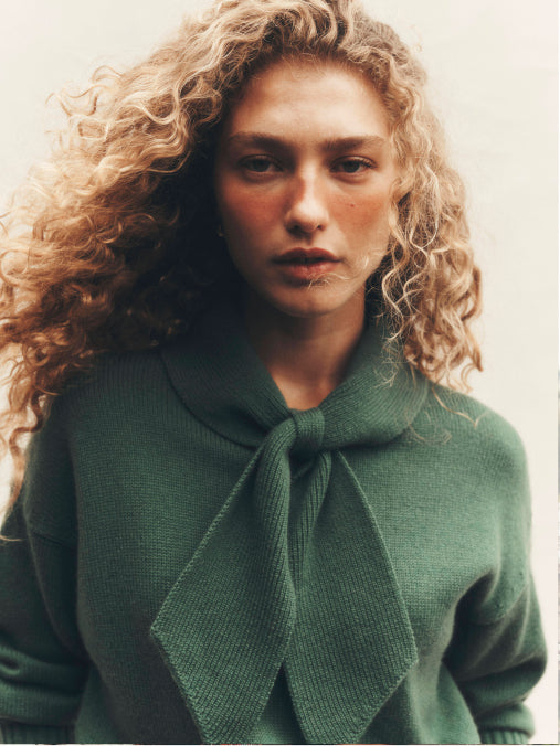 A close-up portrait of a woman with voluminous curly blonde hair wearing a textured dark green knit sweater with a large bow detail at the collar, set against a plain light background.