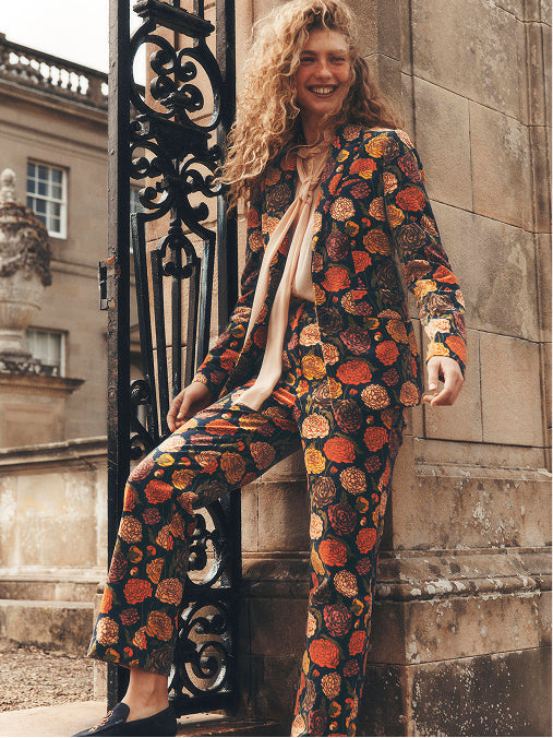 A woman wearing a matching two-piece pajama set with a bold orange, red, and yellow floral pattern on a black background, leaning against a stone wall next to an ornate black metal gate.