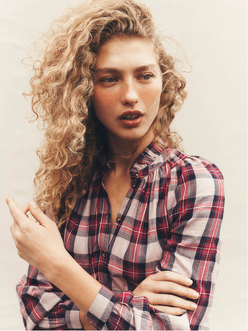 A woman with voluminous curly blonde hair wearing a red, black, and white plaid button-down shirt, posed against a light beige background.
