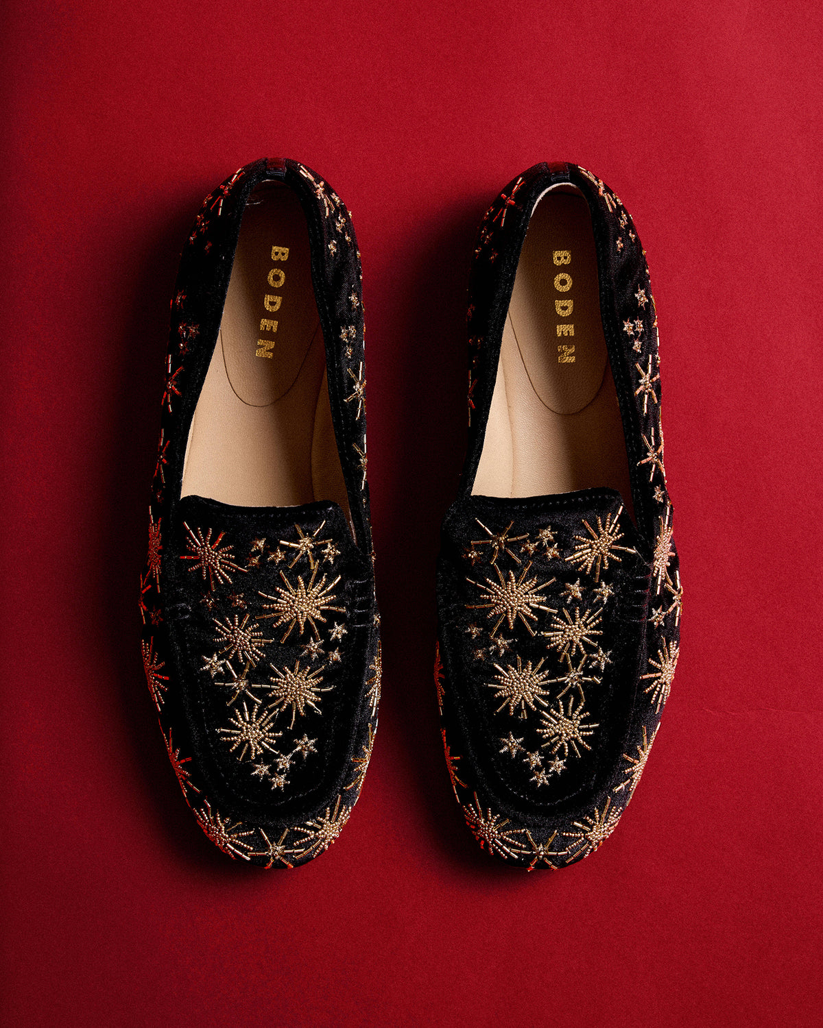 A pair of BODEN slip-on loafers in black velvet with gold embroidered starburst patterns, displayed flat against a solid red background.