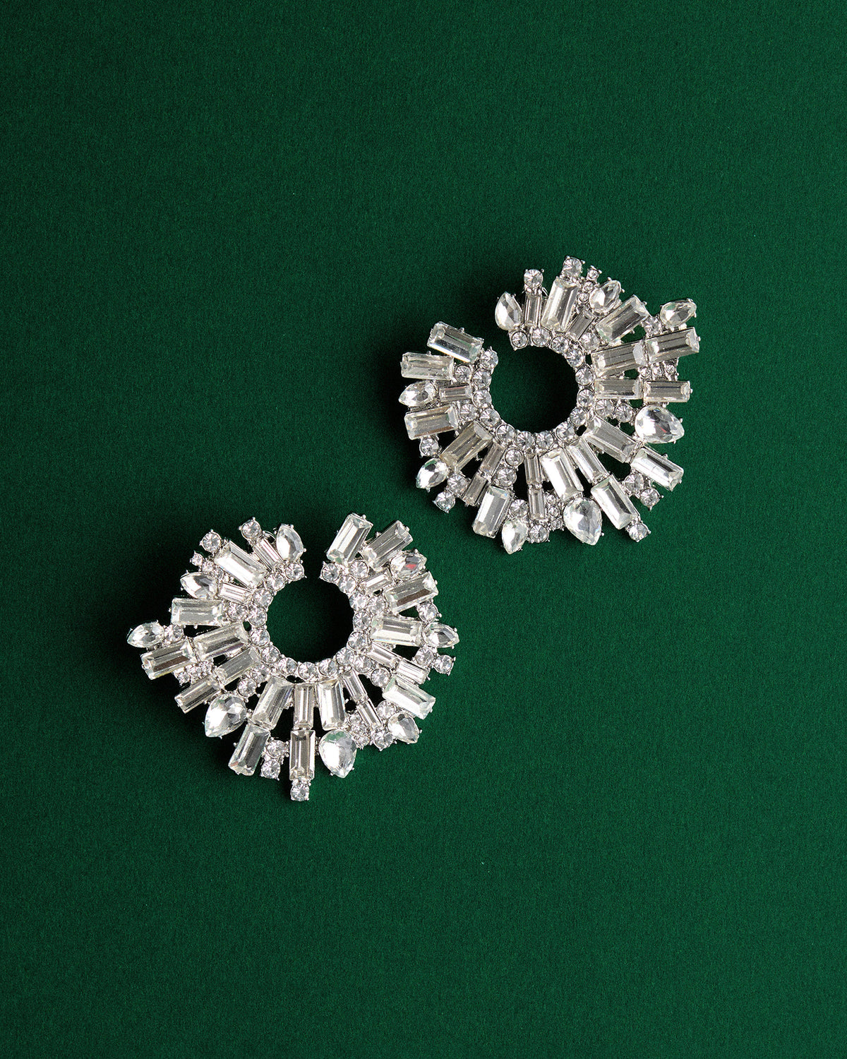 A pair of silver-tone crescent-shaped earrings adorned with clear baguette and round crystals, arranged on a solid dark green background.