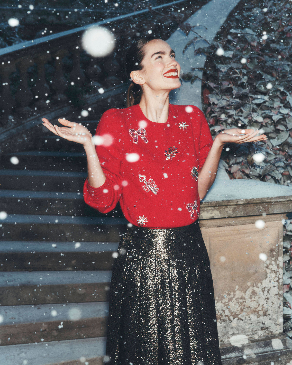 A woman stands outdoors in falling snow, wearing a bright red long-sleeve top with silver embroidered motifs and a black glittery midi skirt.