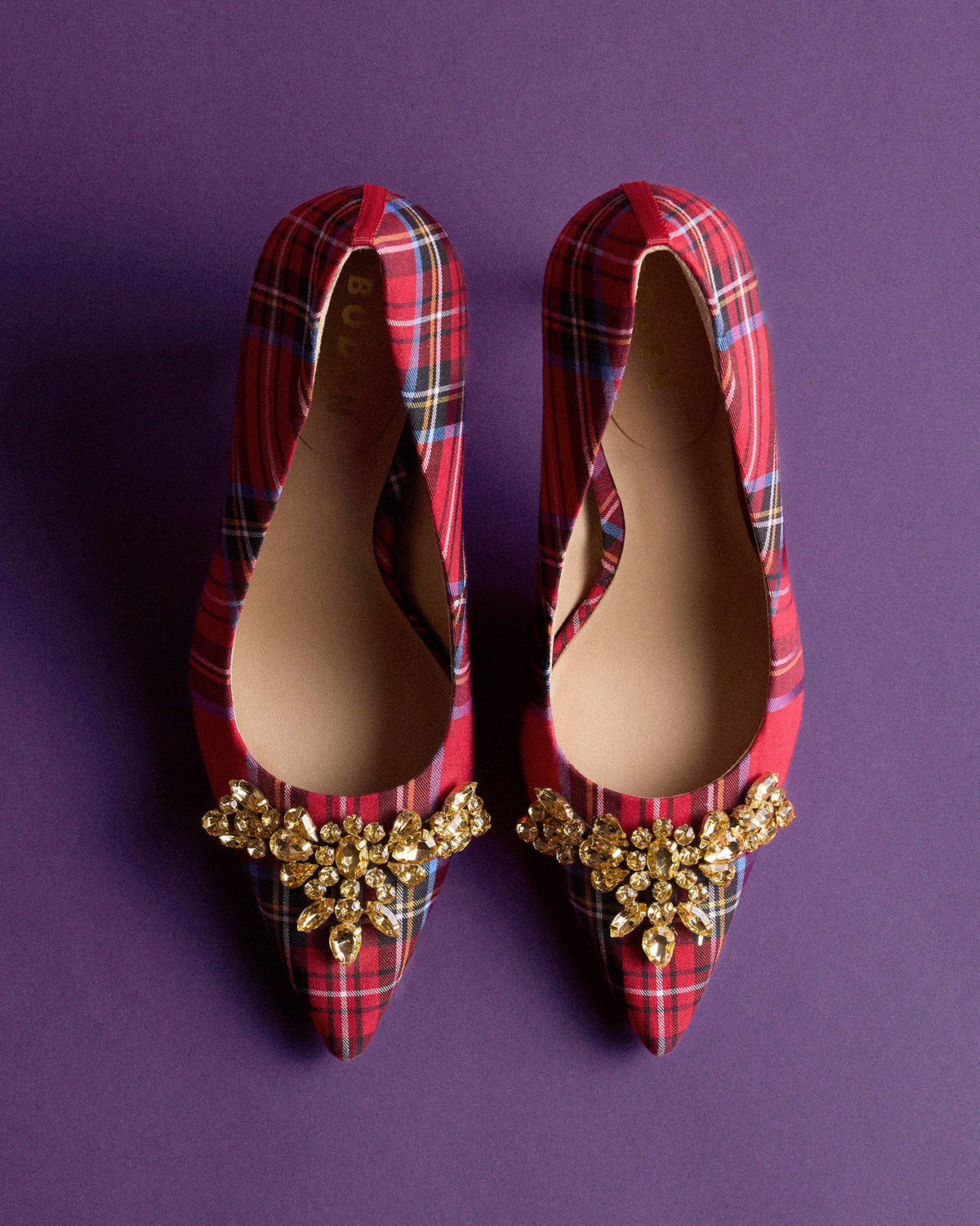 A pair of red and black tartan pointed-toe pumps with ornate gold crystal embellishments on the toes, displayed on a solid purple background.