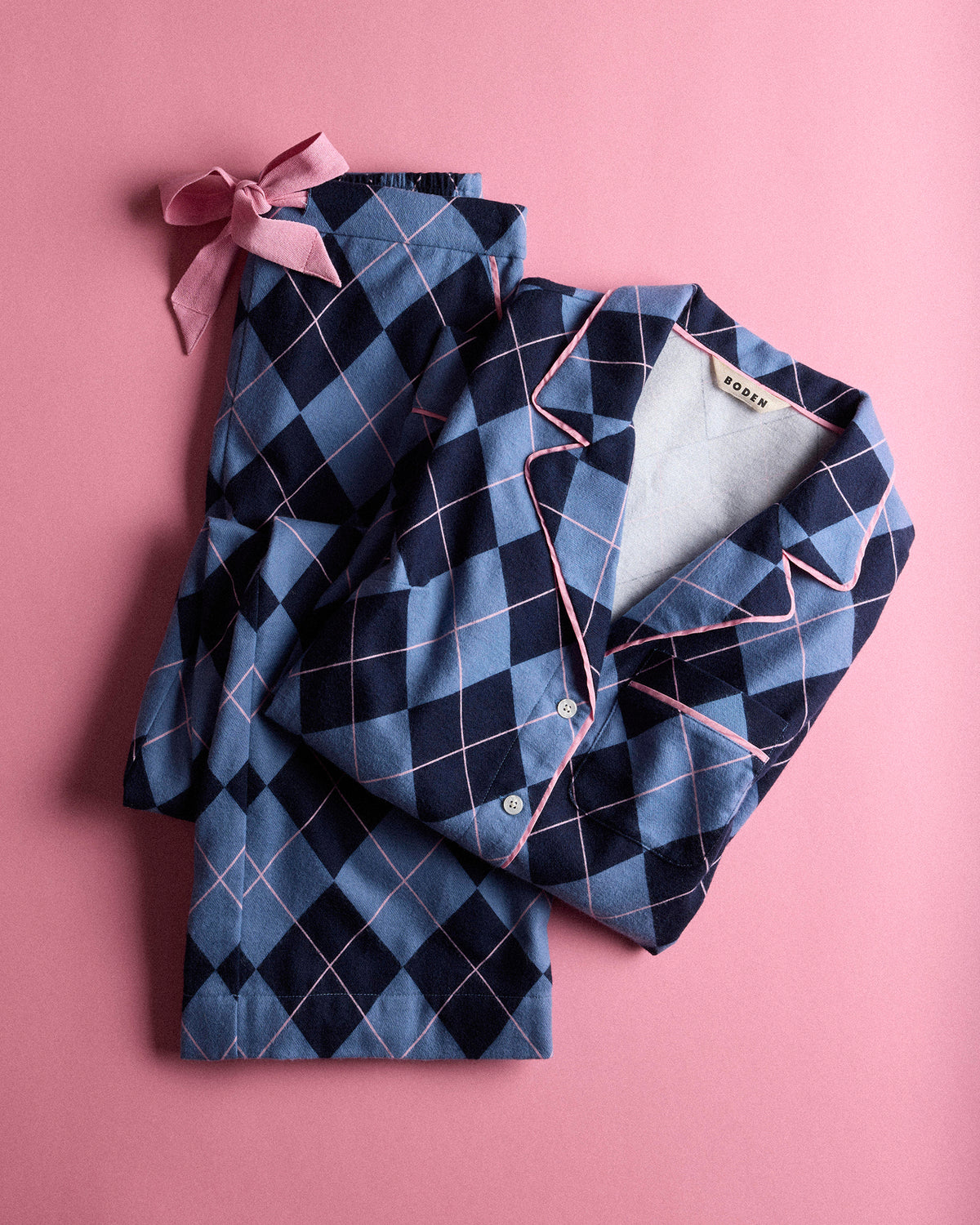 A folded set of pajamas on a solid pink background, featuring a navy and blue argyle pattern with thin pink piping and a matching pink satin bow.