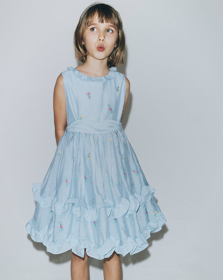 Young girl wearing a light blue dress with floral embroidery on a plain background