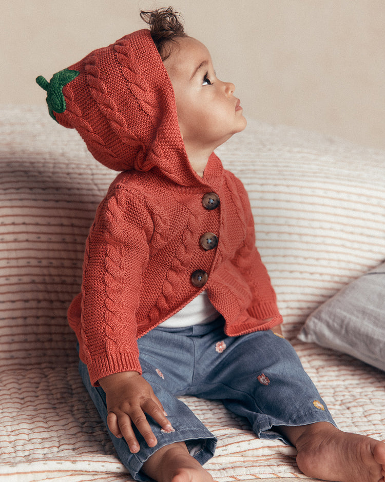 A baby sitting on a striped bed wearing a knitted cowl-neck cardigan and matching hood with a green leaf detail, styled with blue denim pants.