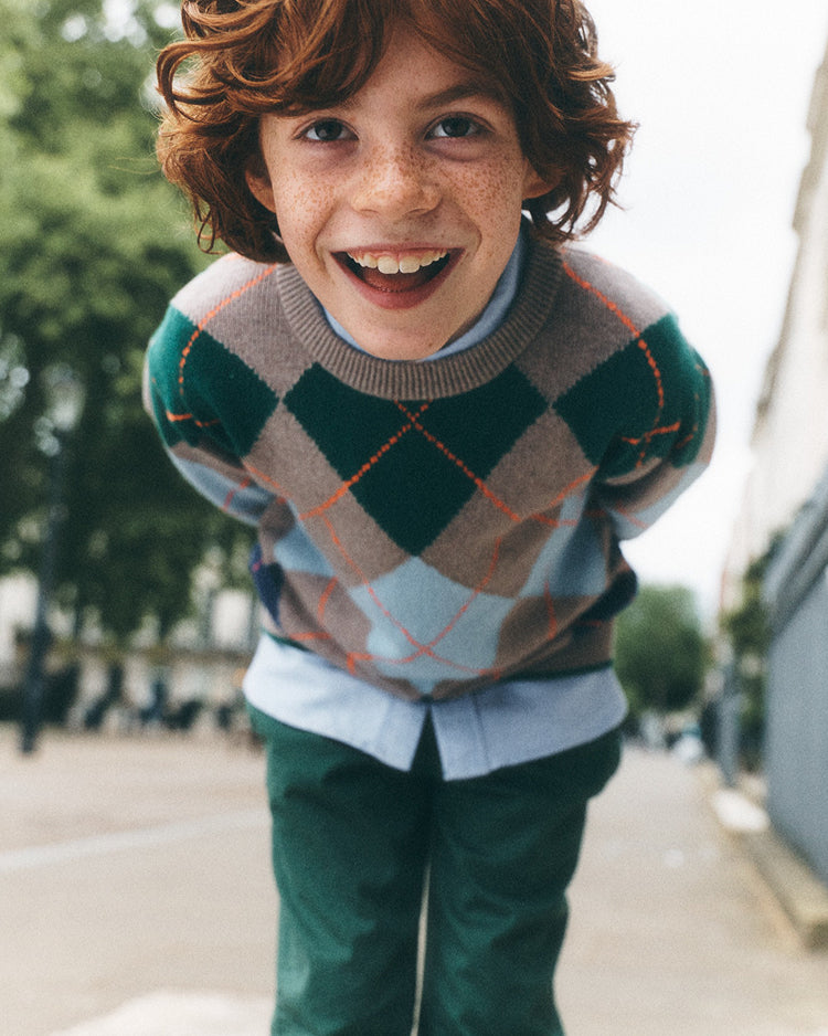 A smiling young boy with curly brown hair and freckles leans forward on a city street, wearing a green, beige, and light blue argyle sweater over a light blue shirt and dark green pants.