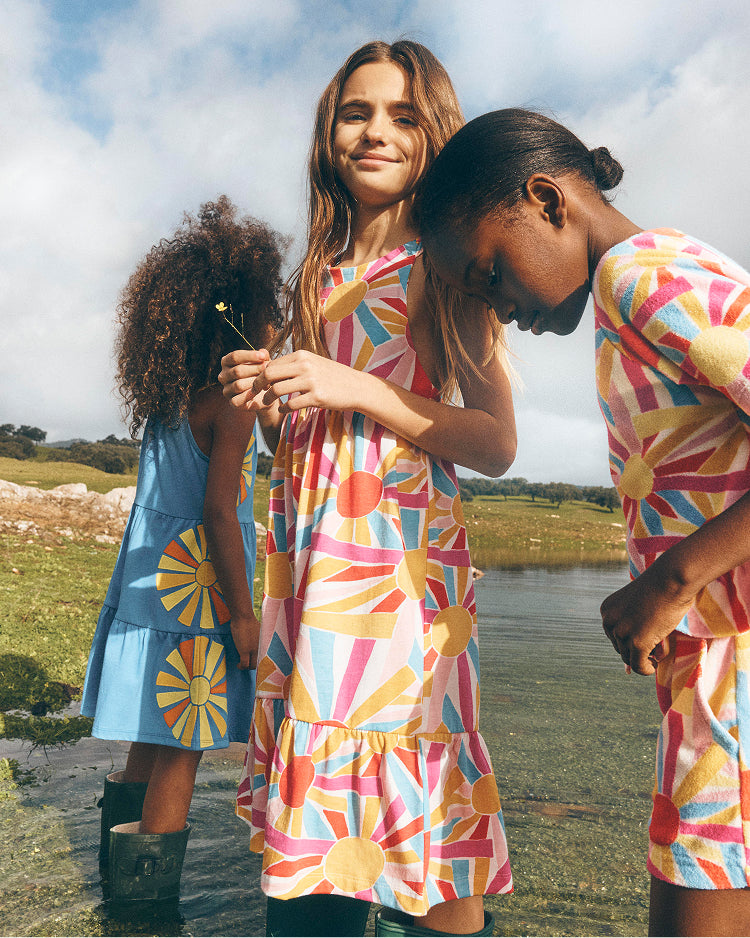 Three young girls in colorful dresses standing by a lake with a scenic background.