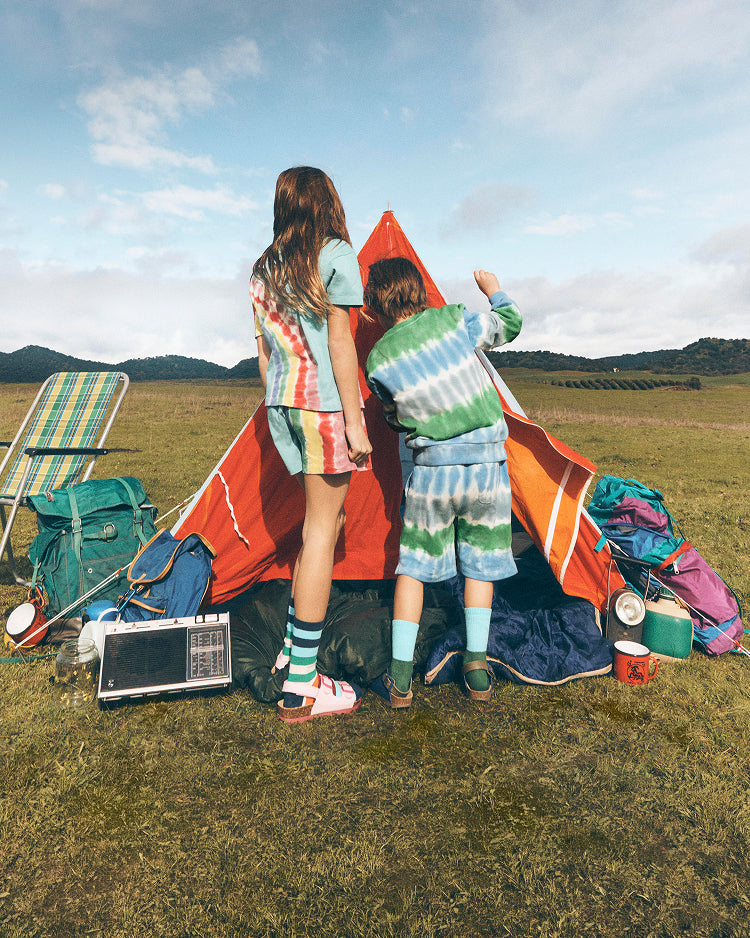 Two children standing next to a colorful tent in a grassy field with mountains in the background.