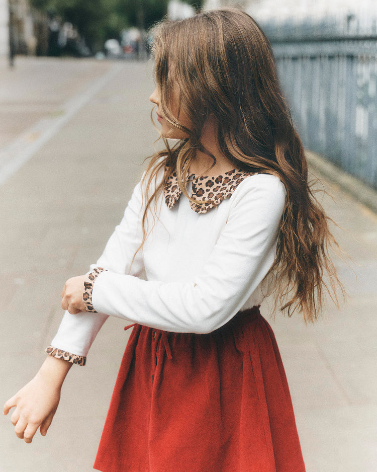 A young girl with long brown hair is shown from a side-back angle outdoors on a sidewalk. She is wearing a white long-sleeve top with a leopard-print collar and cuff, paired with a rust-red skirt.