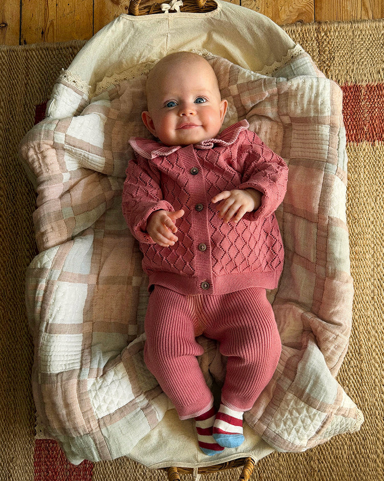 A baby lying on a quilted cream and pink bassinet or bassinet cover, wearing a matching pink cable-knit cardigan and ribbed knit pants with striped socks.