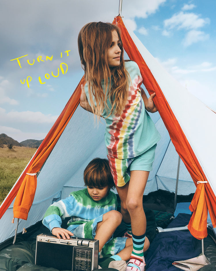 Two children in tie-dye outfits playing with a radio inside a tent with a scenic background.