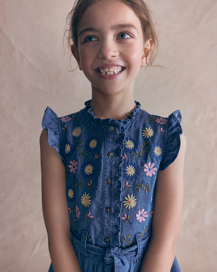 A young girl smiling and looking to the side, wearing a blue denim-style sleeveless top with colorful embroidered flowers and ruffled shoulders, against a plain beige backdrop.