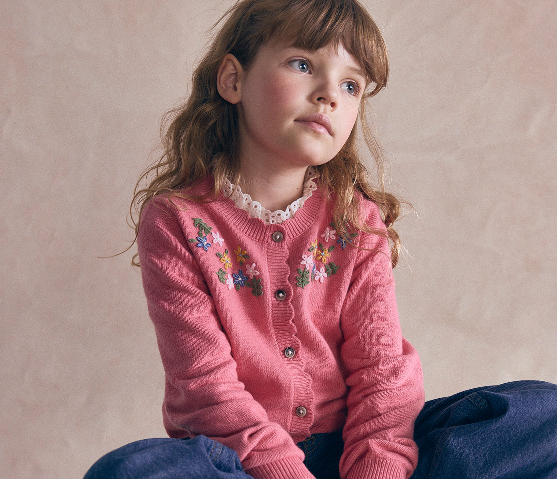 A young girl with light brown hair sits against a textured beige background, wearing a pink button-up cardigan with colorful floral embroidery on the chest and dark blue pants.