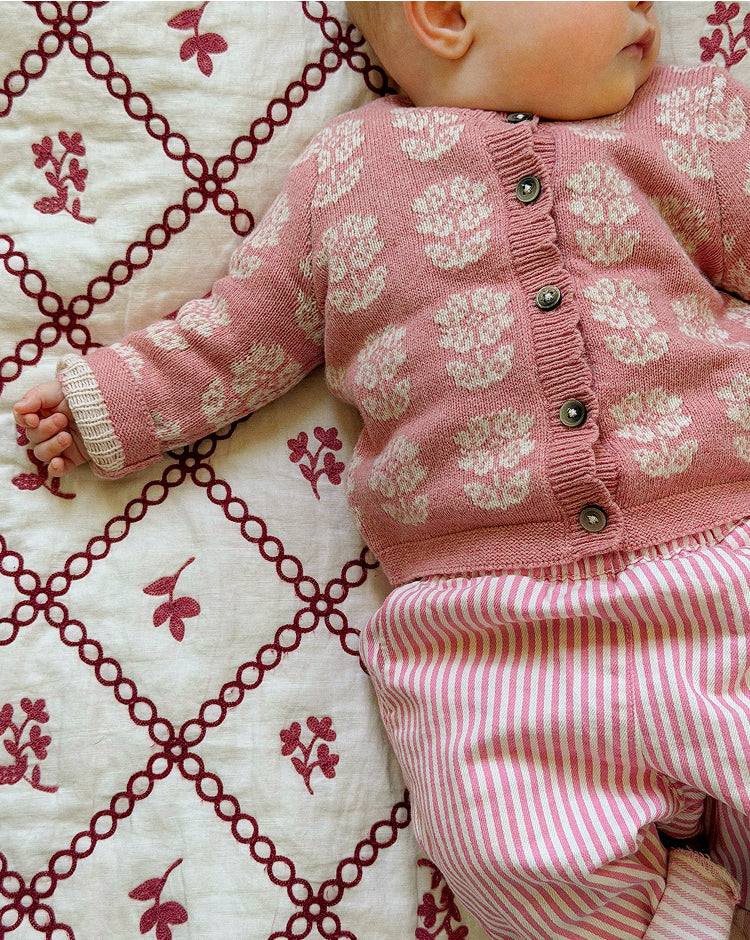 A close-up of a baby lying on a cream quilt with a red floral lattice pattern, wearing a pink knit cardigan with cream floral motifs and pink-and-white striped pants.