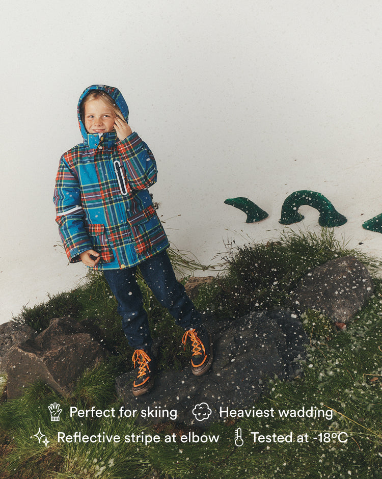 A child wearing a blue, red, and orange plaid hooded ski jacket, dark pants, and orange shoes stands on rocks outdoors against a white wall, with text overlays highlighting product features.