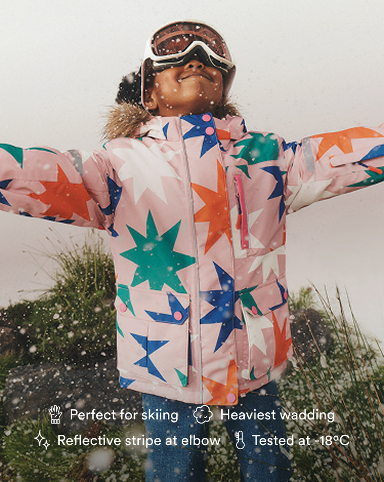 A child wearing a light pink ski jacket with a colorful star pattern, goggles, and a fur-trimmed hood, standing outdoors in the snow with arms outstretched. Text overlays at the bottom highlight product features.