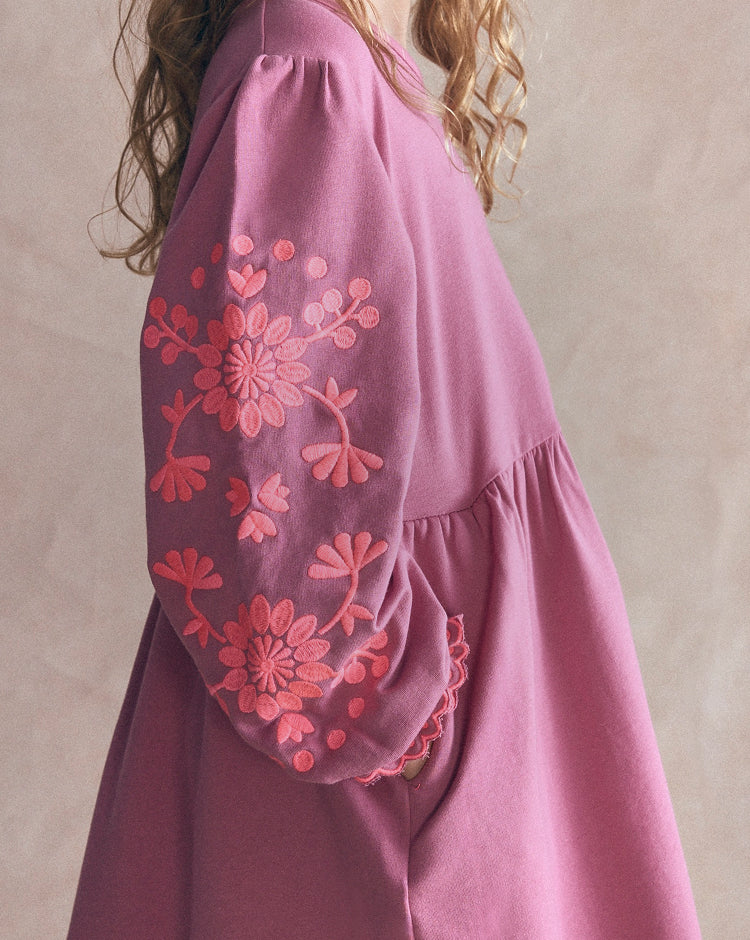 A close-up of a dusty pink dress with voluminous sleeves featuring pink floral embroidery, worn by a model against a neutral beige background.