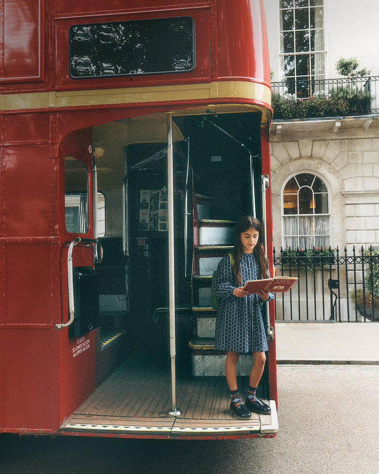 A young girl in a blue patterned dress stands in the doorway of a red double-decker bus, holding a book. The photo is taken on a street with a classic stone building in the background.