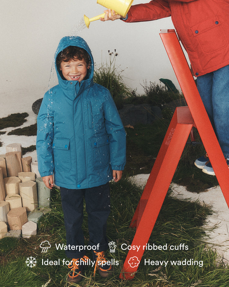 A child wearing a bright teal waterproof hooded jacket stands on grass while water is poured over the shoulders from above by an adult in a red jacket on a red step ladder. Product text overlays at the bottom mention waterproof, cosy ribbed cuffs, ideal for chilly spells, and heavy wading.