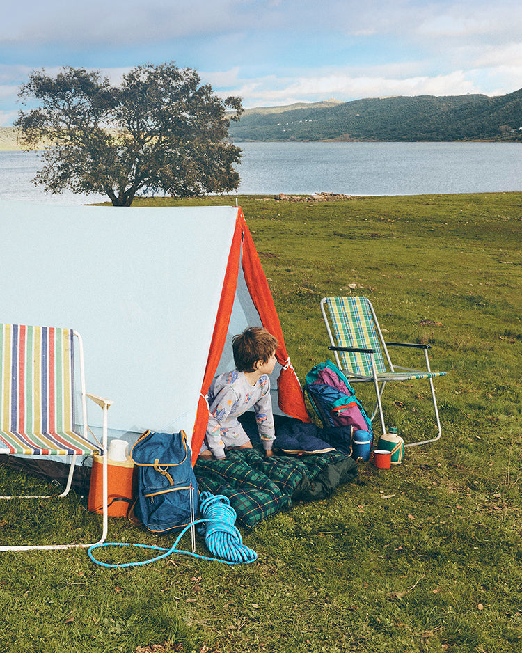Child playing near a red tent with camping gear on a grassy area by a lake.