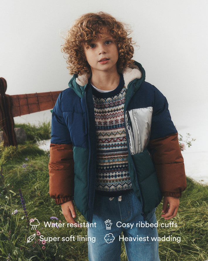 A child wearing a color-blocked puffer jacket outdoors on grassy ground. The jacket is navy blue on top, teal green on the lower front, and brown on the sleeves, with a silver zipper and a patterned sweater underneath.