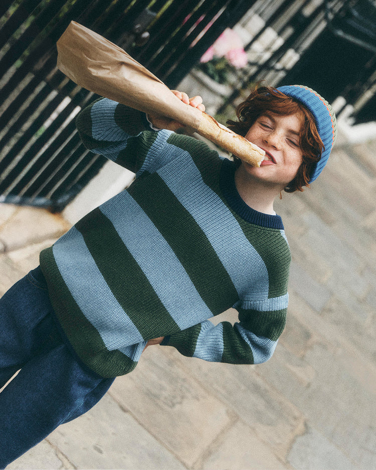 A child wearing a green and light blue striped knit sweater and a blue beanie hat, holding a baguette outdoors on a stone pavement.