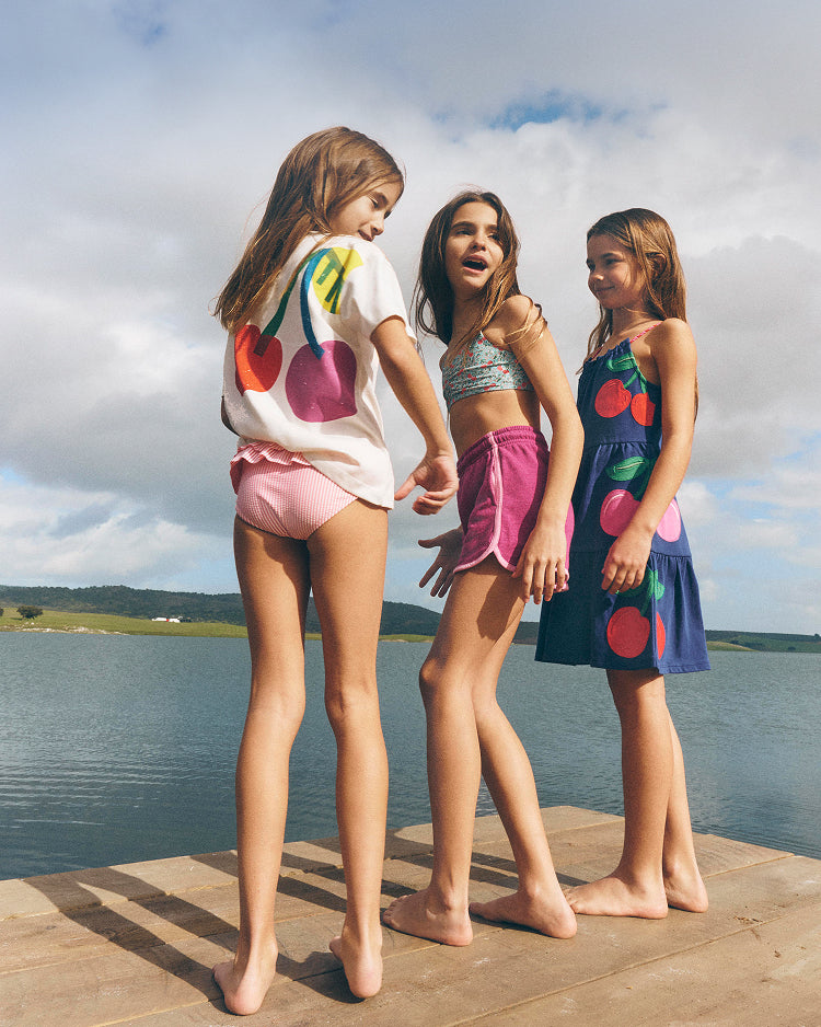 Three young girls standing on a dock by a lake, wearing cherry-themed clothing.