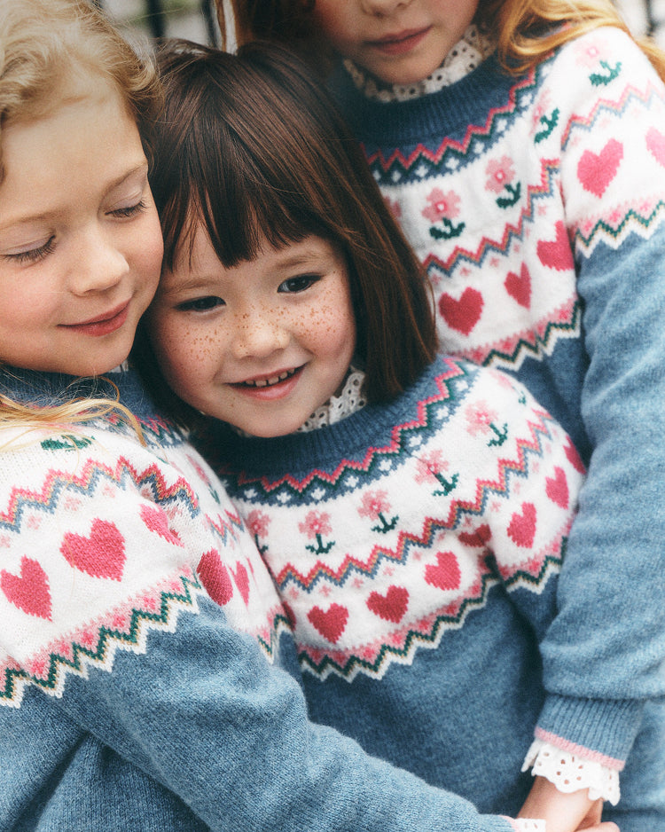 Three children wearing matching blue knit sweaters with a white Fair Isle pattern featuring pink hearts, red zigzags, and small floral motifs.