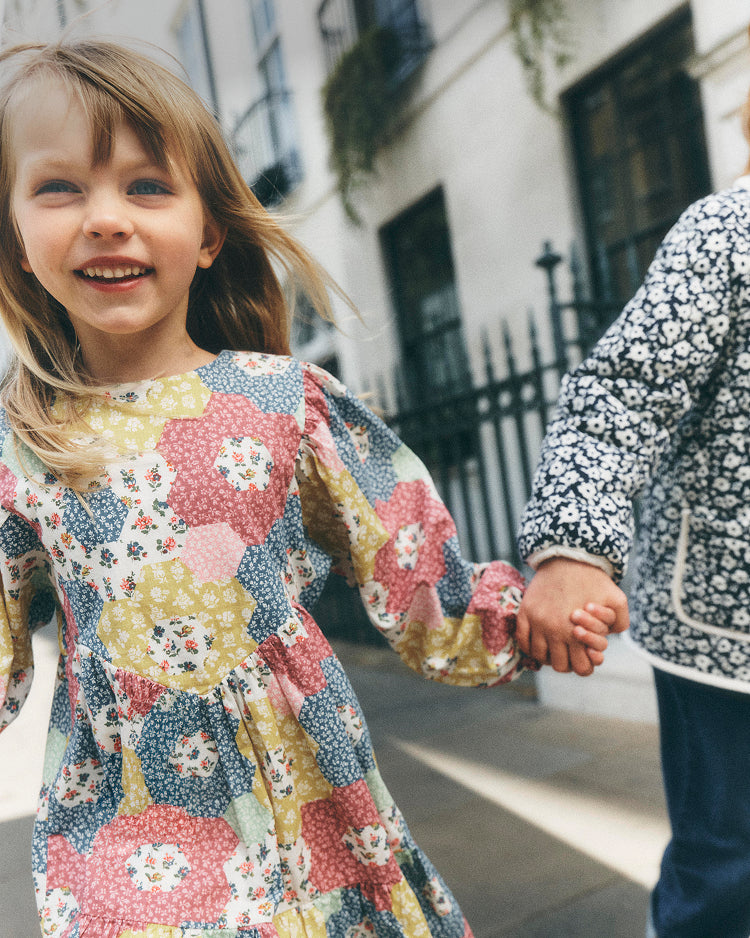 A young girl smiling and holding hands with another child outdoors. The girl wears a long-sleeve dress with a colorful floral and geometric patchwork pattern in pink, yellow, blue, and white.