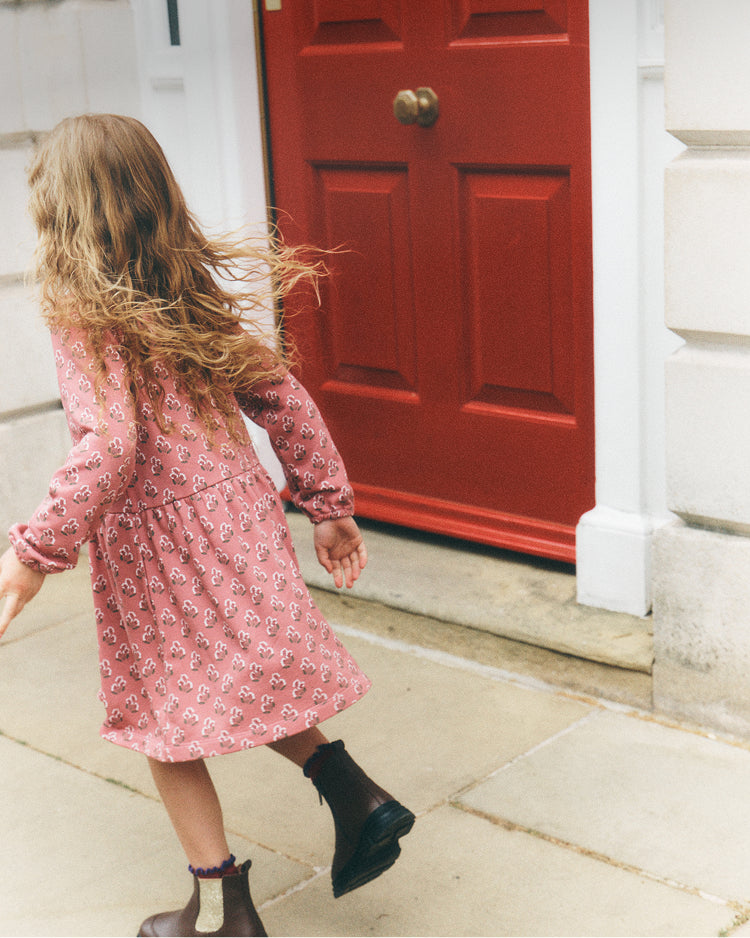 A young girl with long blonde hair is seen from behind, wearing a pink floral print dress and dark boots while walking on a stone sidewalk in front of a red door.