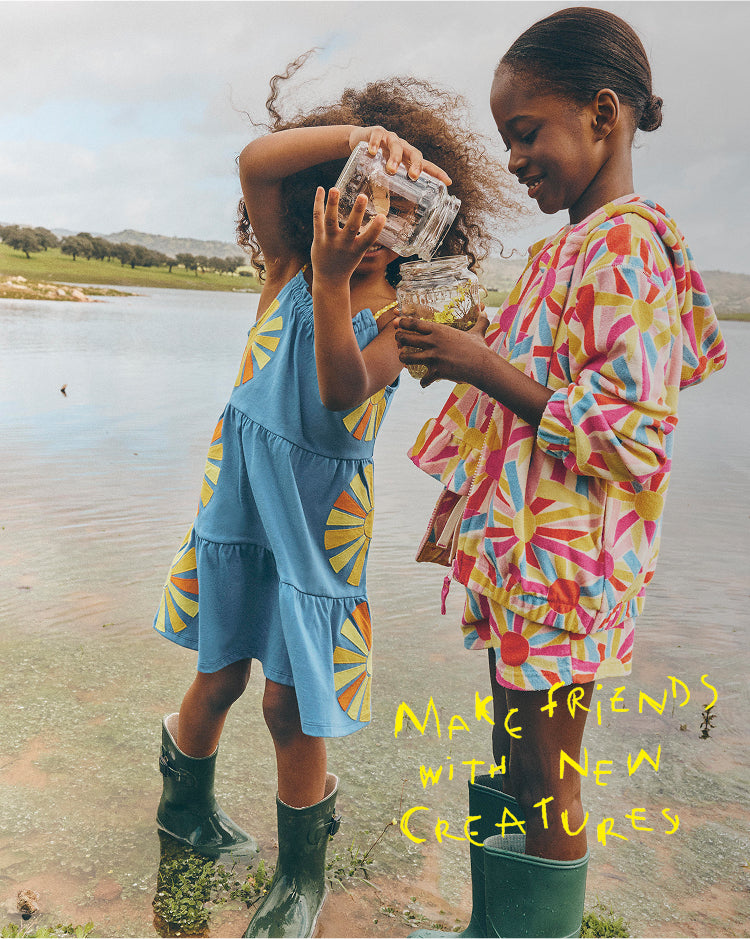 Two children by a lake, one holding a jar with a small creature, wearing colorful raincoats and rain boots.