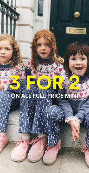 Three young girls sitting on stone steps in front of a dark door, wearing matching knit sweaters and patterned navy pants. Large yellow text across the center reads '3 FOR 2 ON ALL FULL PRICE MINI'.