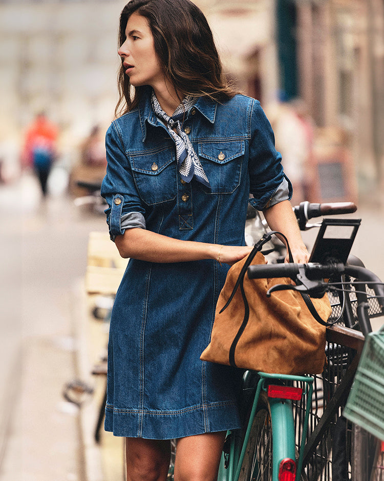 A woman wearing a blue denim shirt dress stands on a city street next to a green bicycle, holding a tan suede shoulder bag.