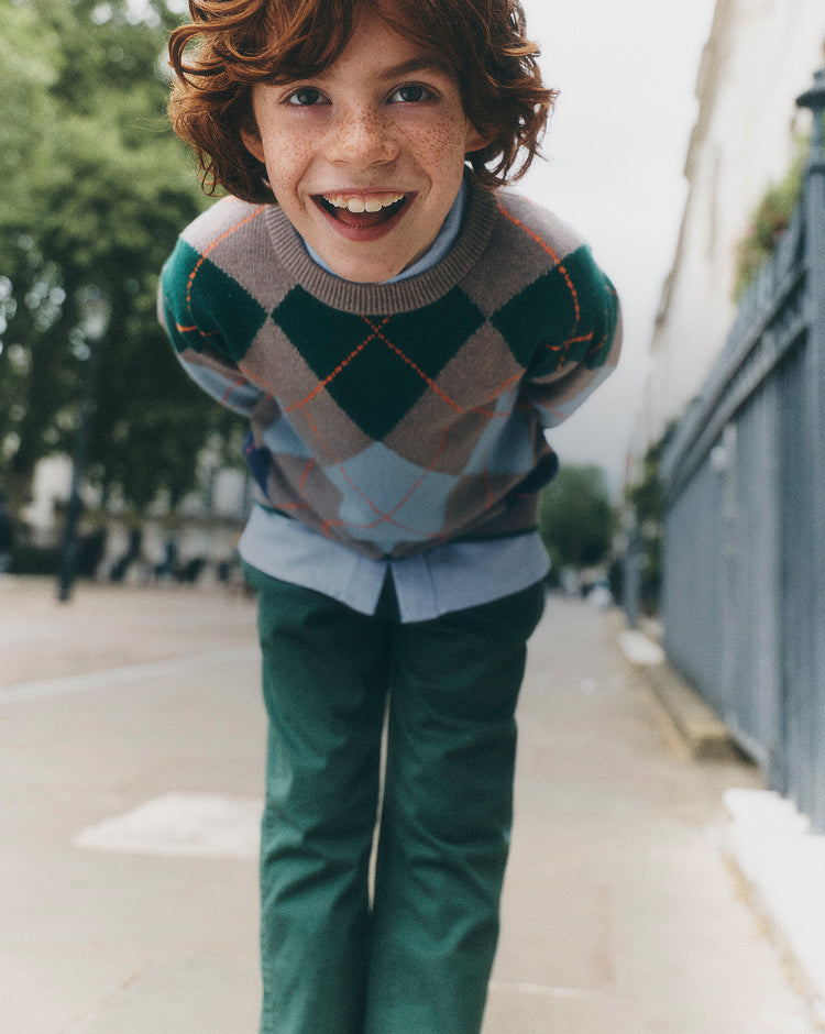 A smiling young boy with curly brown hair and freckles leans forward on a city sidewalk, wearing a green, gray, and light blue argyle sweater over a light blue shirt, paired with dark green trousers.