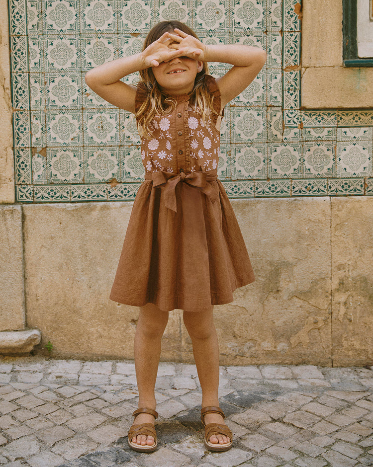 Young girl in a brown dress with floral embroidery standing in front of a tiled wall.