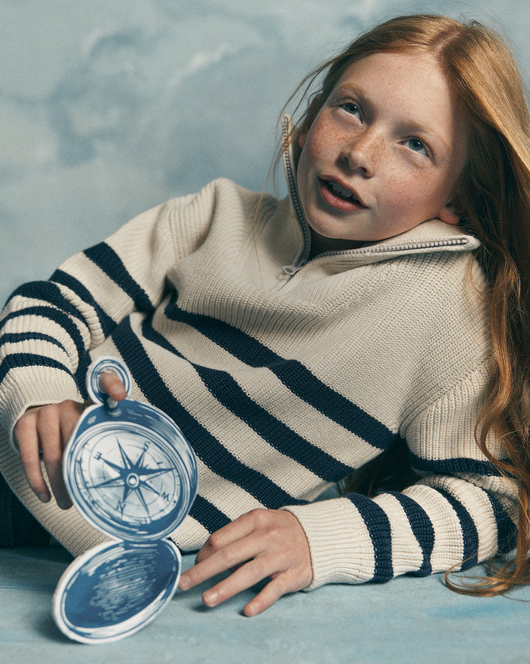 A young girl with red hair lies on a blue surface wearing a cream and navy striped quarter-zip sweater, holding a blue-and-white compass.