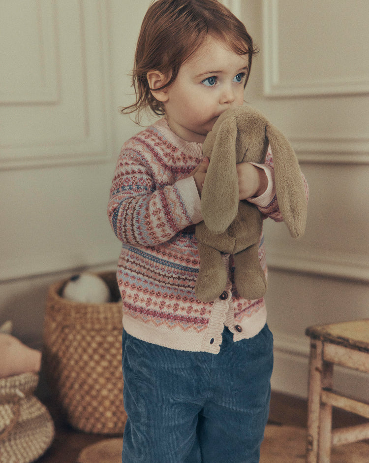 A toddler stands indoors holding a brown plush bunny. The child is wearing a Fair Isle knit cardigan with pink, blue, and cream patterns, paired with blue corduroy pants.