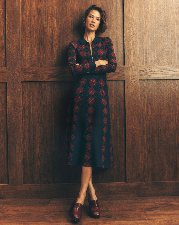 A woman stands against a dark wood-paneled wall wearing a long-sleeve midi dress with a red and black argyle pattern, paired with dark brown leather loafers.