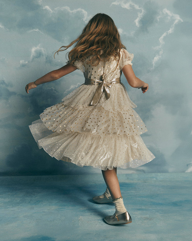 A young girl with long light brown hair is shown from behind, wearing a cream-colored, tiered tulle dress with gold polka dots and a matching ribbon belt. She is posing in front of a blue, cloud-textured backdrop.