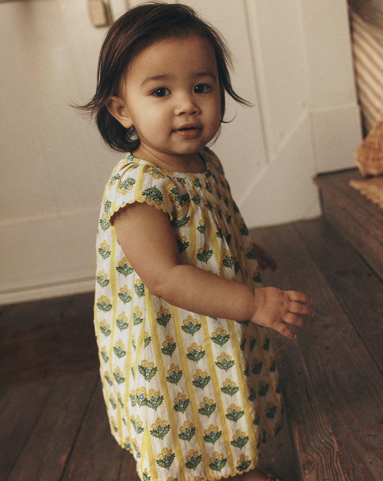 Child in a yellow and green patterned dress standing on a wooden floor.