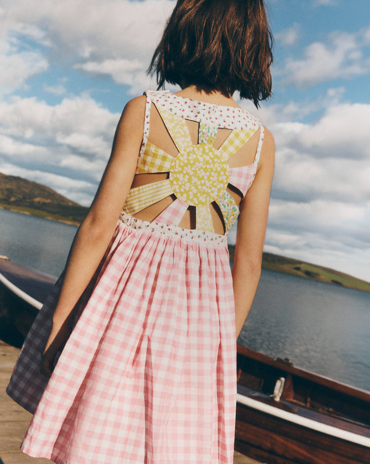 Person wearing a pink checkered dress with a sun-shaped cutout back, standing on a boat with a scenic background.