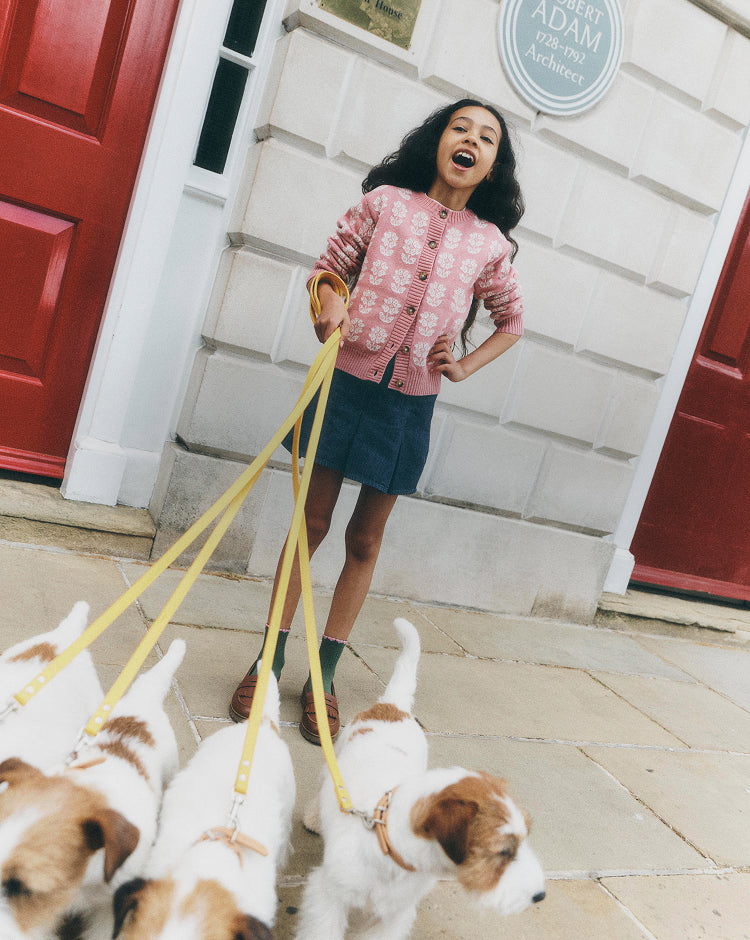 A young girl laughing while walking four small dogs on yellow leashes in front of a white building with red doors. She is wearing a pink patterned cardigan, a dark blue skirt, and green socks.