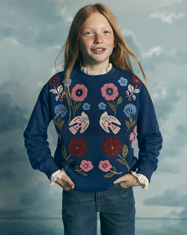 A young girl with long strawberry-blonde hair stands against a cloudy blue-gray sky backdrop, wearing a navy blue crewneck sweater decorated with colorful floral and butterfly embroidery, paired with blue jeans.