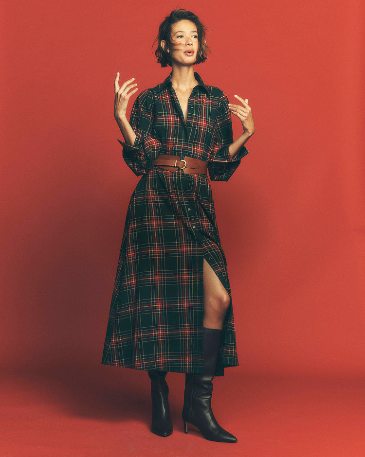 A woman stands against a solid red studio background wearing a long-sleeve plaid shirt dress in black, red, and green, styled with a brown belt and black knee-high boots.