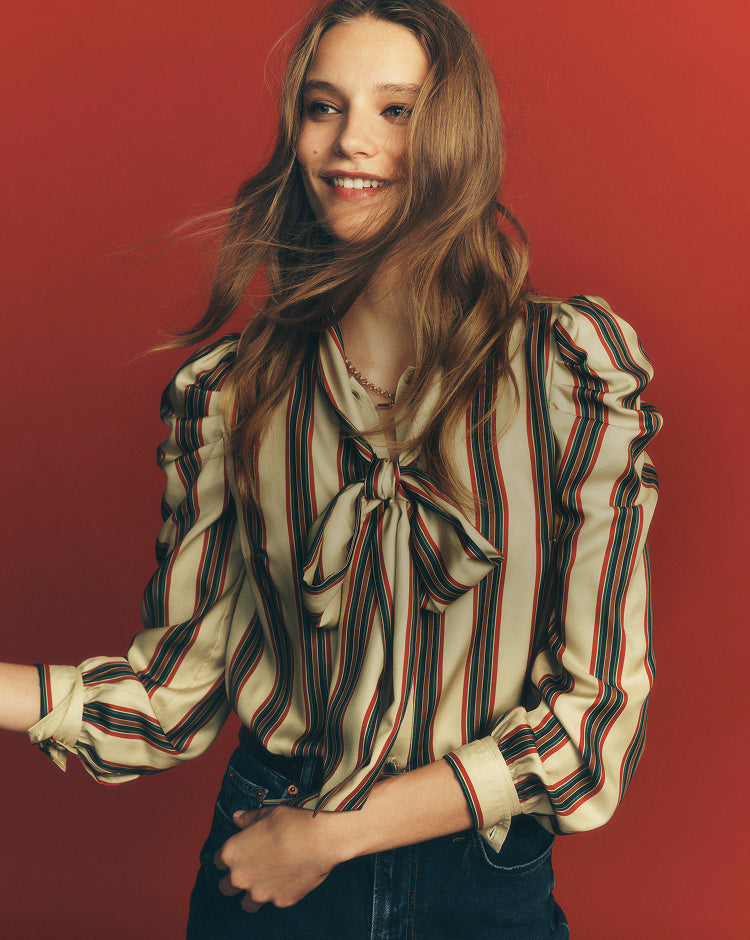 A woman with long light brown hair wearing a cream-colored blouse with vertical red and green stripes and a front bow tie, paired with dark blue jeans, posed against a solid red background.